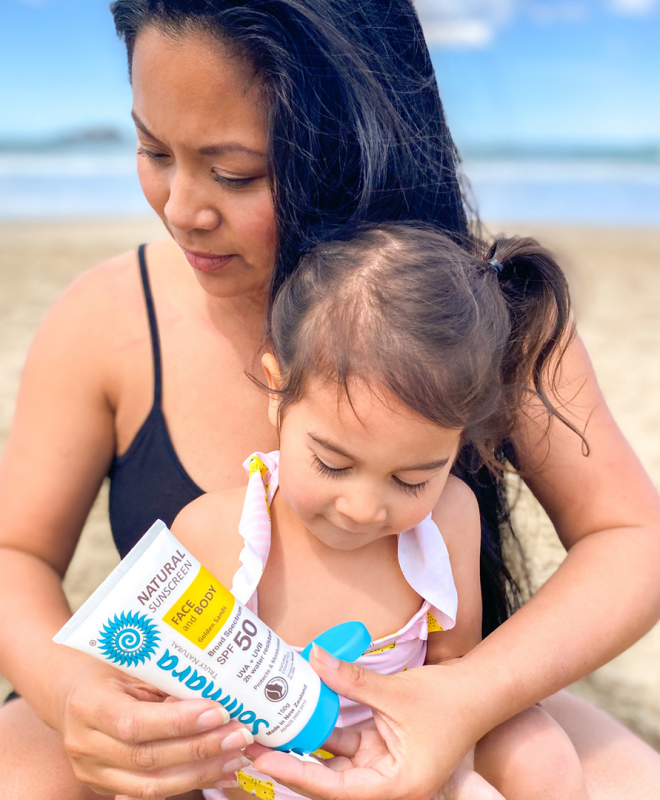 Mother and young daughter on a sandy beach applying sunscreen together from a tube labeled SPF 50+ sunscreen.