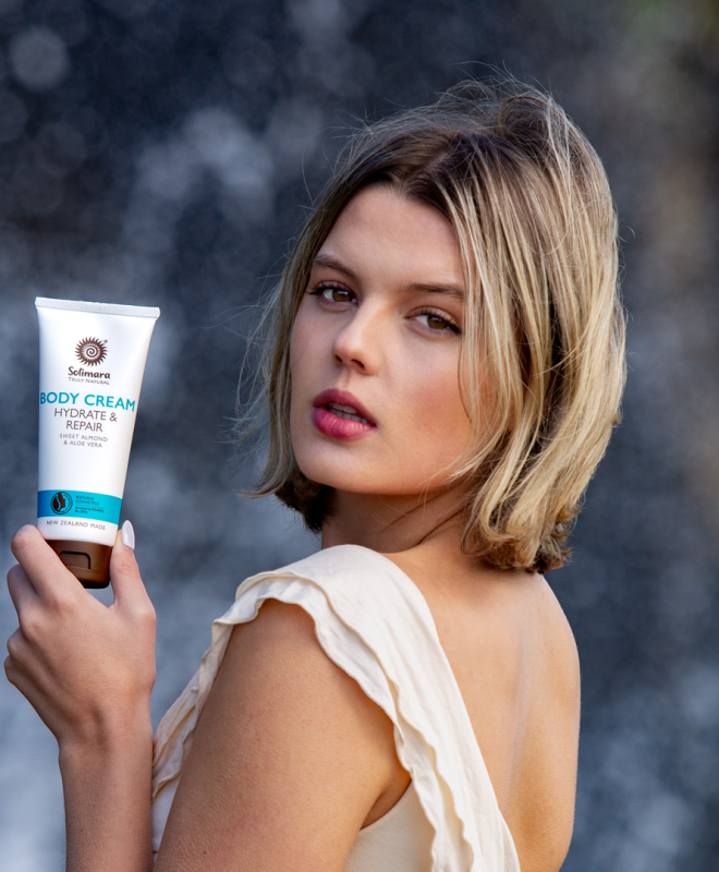 Young woman holding a white tube of Solimara Body Cream Hydrate & Repair, posed outdoors with a blurred dark background.