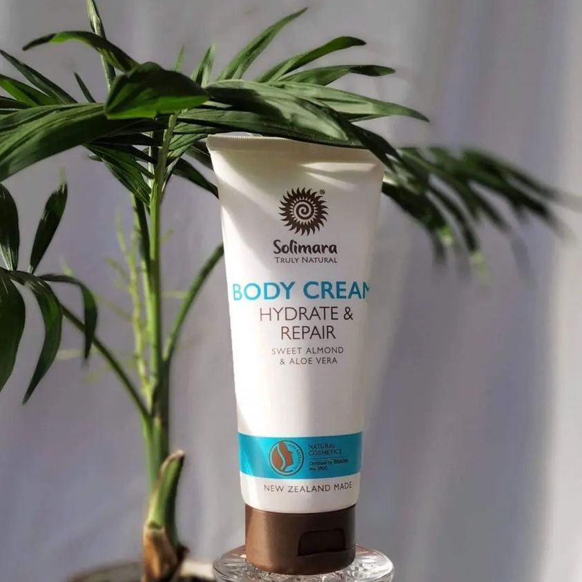 Solimara Body Cream tube labeled 'Hydrate & Repair' with sweet almond and aloe vera, standing on a glass pedestal beside a plant.